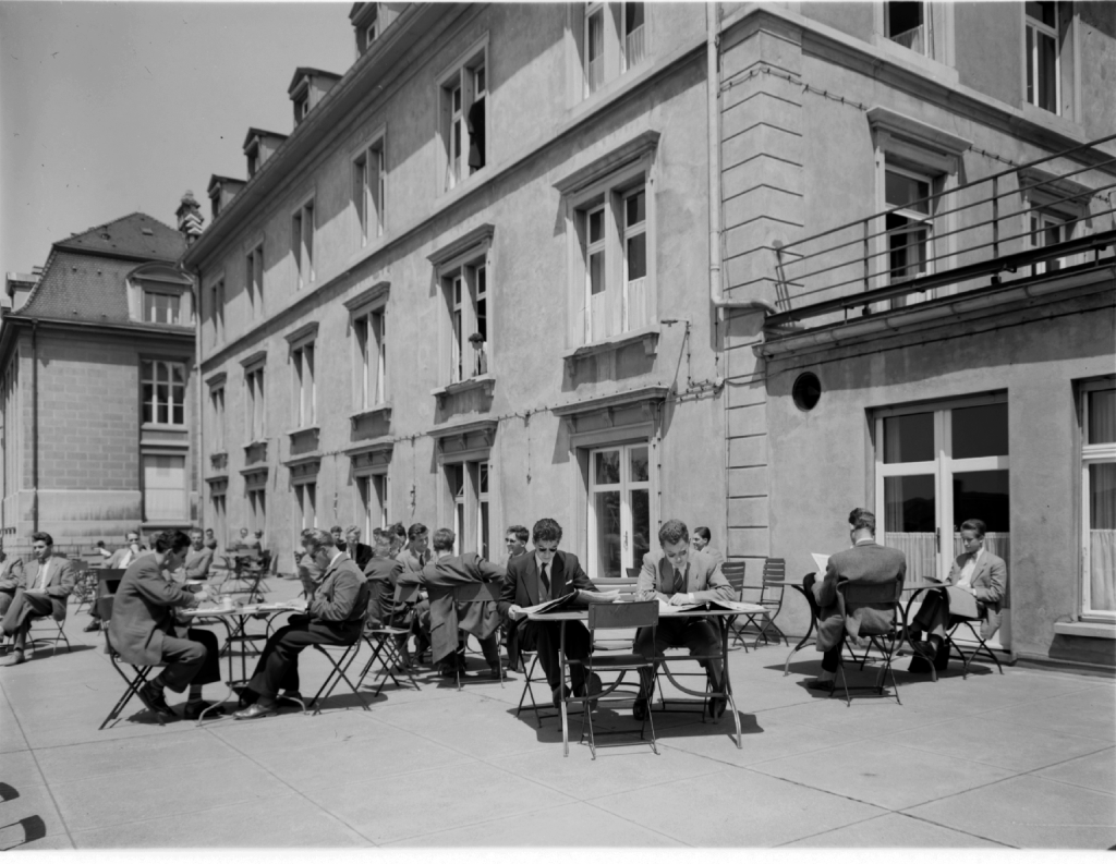 Black and white picture of students sitting on a terrace in front of a house. They are writing and reading in the sun.