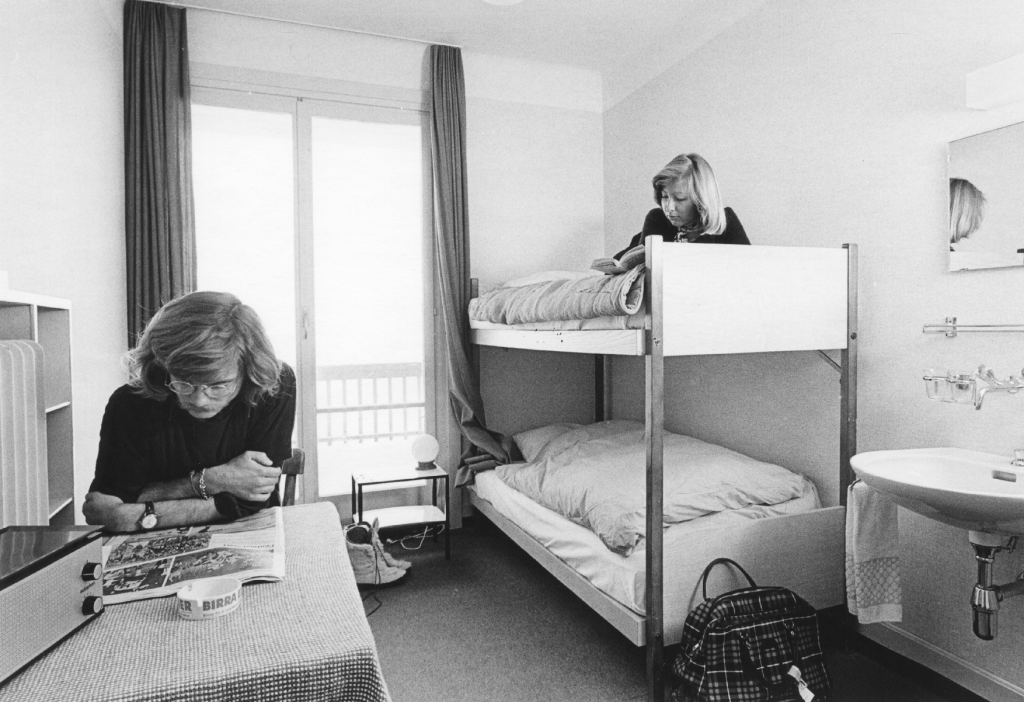 Two students in a hotel/hostel room. Both are reading. One is on the table and the other on a bunkbed.